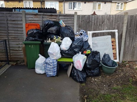 A collection of various rubbish bags and containers stacked against a wooden garden fence in a residential outdoor area. The black rubbish bags, some tied and others loosely closed, are piled on top of a green wheelie bin and a small green plastic container with a lid. Additional white and blue striped bags are stacked on a small wooden bench positioned next to the bins. Behind the bags, there is a white-painted window frame leaning against the fence. The ground is paved with asphalt, with a patch of soil and some grass visible on the right side of the image. This scene appears to be part of a garden clearance or rubbish removal effort, typical of services provided by companies like Garden Clearance Newham, serving local residents in Newham postcode areas.