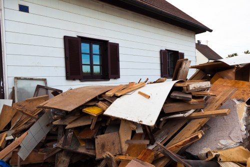 A large pile of assorted discarded wooden planks, panels, and debris stacked haphazardly in front of a white residential building with dark brown window shutters, likely created during a garden clearance or renovation project near Newham, suggesting the need for professional rubbish removal services such as those offered by Garden Clearance Newham. The wooden materials vary in size, colour, and texture, with some pieces showing rough, splintered edges and weathered surfaces, while others are smoother and painted or finished in shades of brown and white. The pile extends across a paved or gravel surface, partially obstructing the view of the building's exterior, which features two closed windows with lattice-style panes. The scene is set in daylight, indicating typical outdoor conditions, with no visible activity or personnel present. The context aligns with waste management services that handle garden or construction rubbish in the Newham area, emphasizing the need for effective debris clearance to maintain property tidiness.