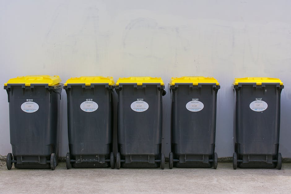A close-up view of a pile of crushed aluminum cans and plastic bottles, primarily consisting of soft drink containers in various colors such as silver, red, blue, black, and yellow. The cans are tight